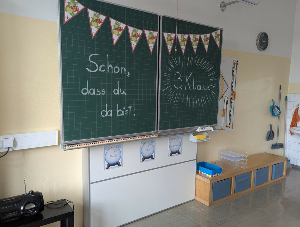 Eine grüne beschriftete Tafel in der Klasse. Darauf steht "Schön, dass du da bist!"