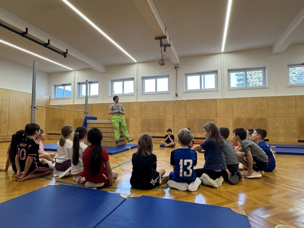 Turnunterricht: Kinder sitzen im Kreis im Turnsaal. Stationen werden erklärt. Rundherum liegen blaue Matten. 