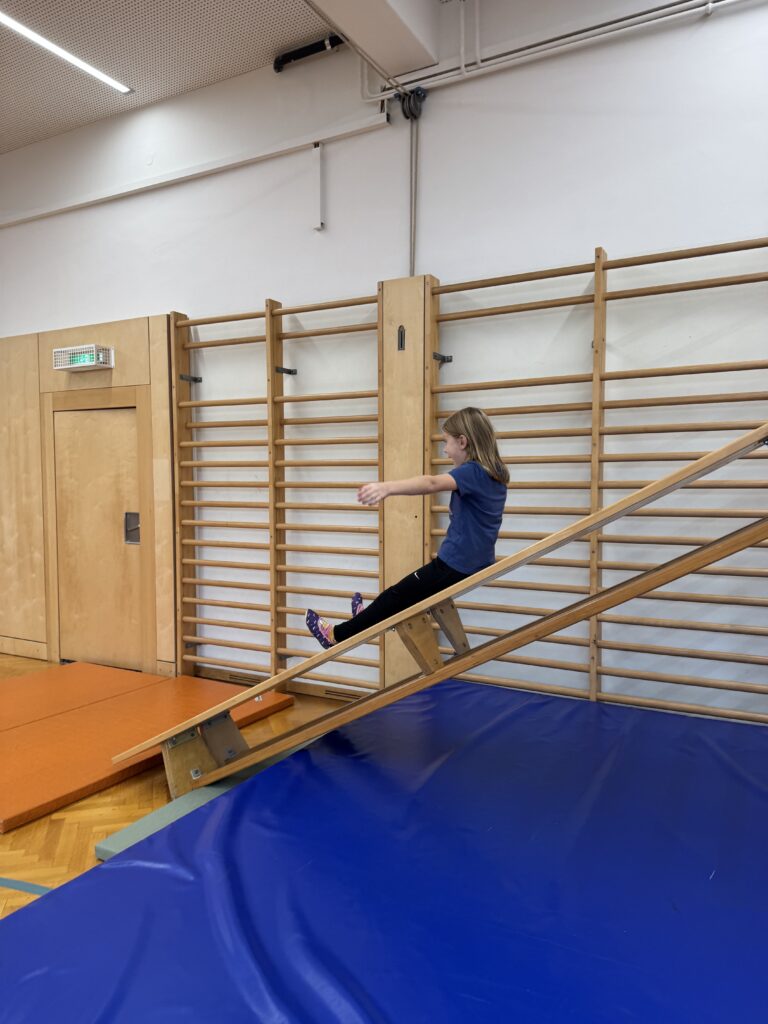 Im Turnsaal sind Stationen vorbereitet. Ein Kind rutscht von einer schrägen Bank auf eine Matte. 