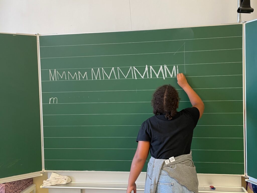 An der grünen Tafel schreibt ein Kind den Buchstaben "M".