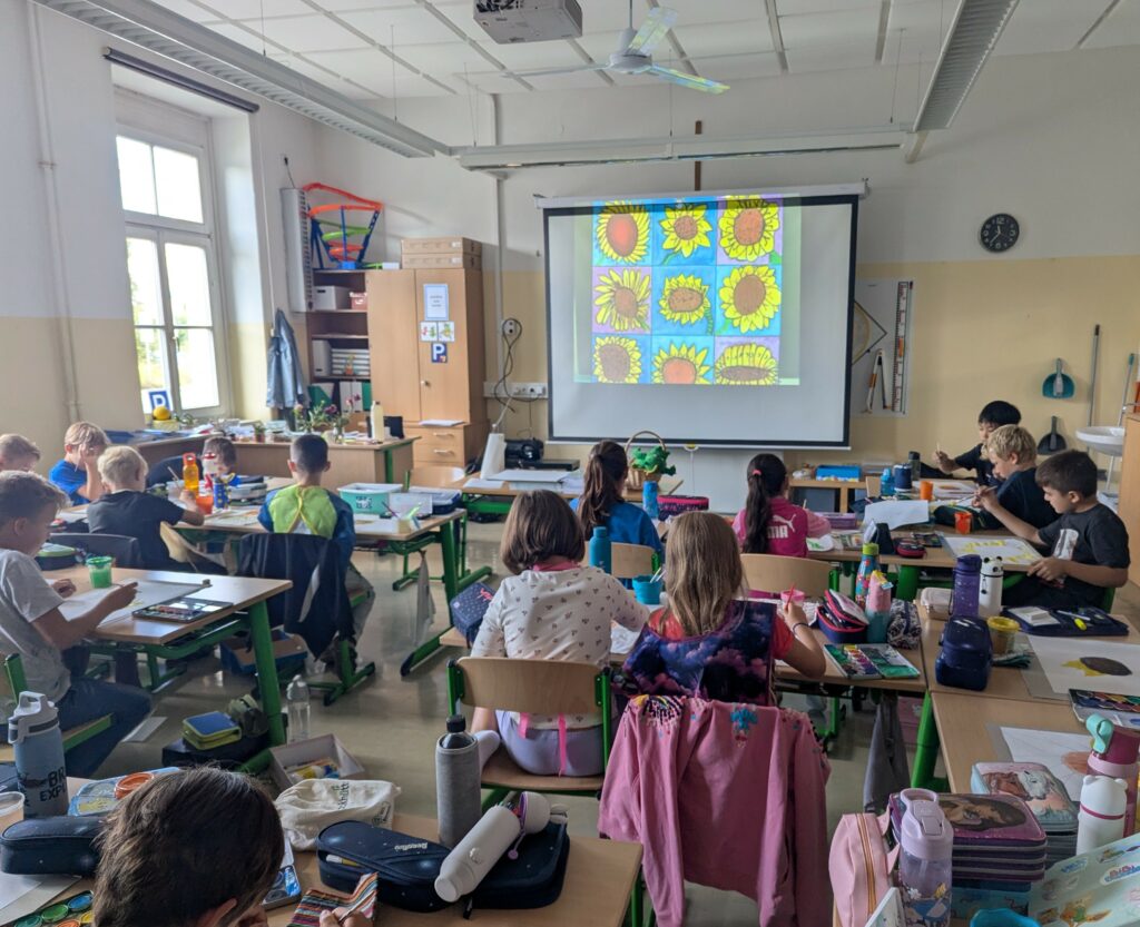 Kinder sitzen in der Klasse und über den Beamer kann man die wunderschön gestalteten Sonnenblumen der Kinder sehen. 