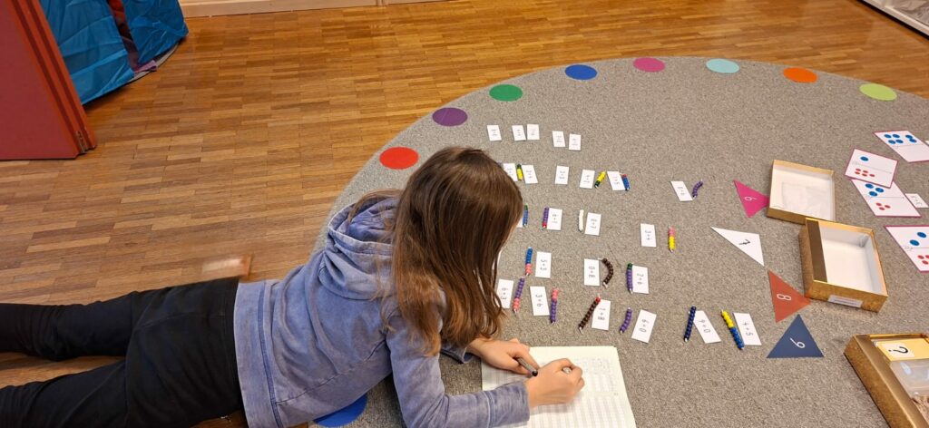 Ein Kind sitzt am Teppich und arbeitet mit den "Goldenen Schachteln". Dort werden Rechnungen mit dem bunten Perlenmaterial gelegt. 