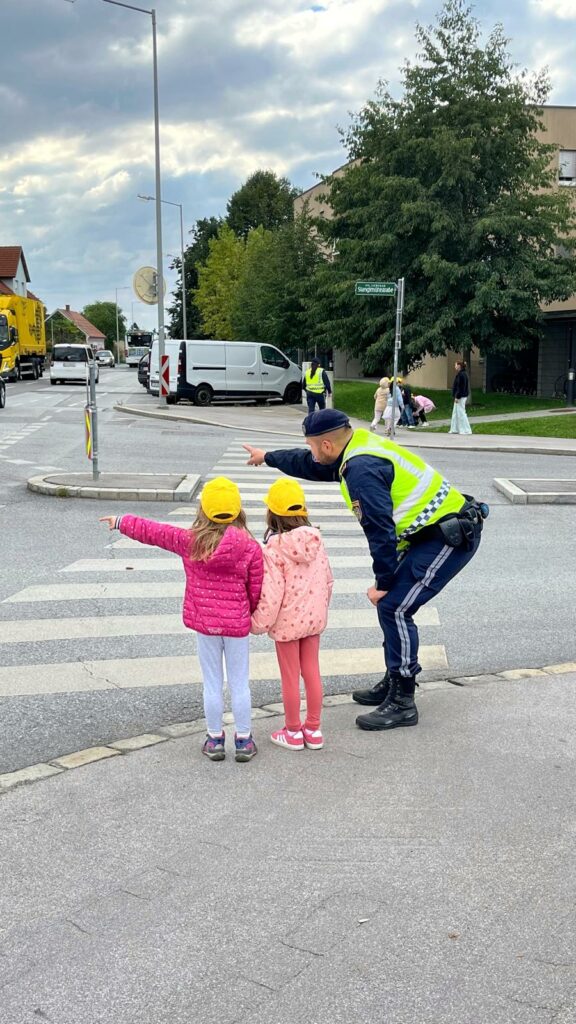 "Verkehrserziehung": Zwei Kinder mit gelben Kappen stehen vor dem Zebrastreifen mit dem Polizisten und wollen die Straße überqueren. 